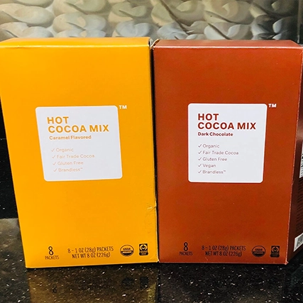 Two boxes of hot cocoa mix on a black countertop. One box is caramel-flavored, and the other is dark chocolate-flavored.